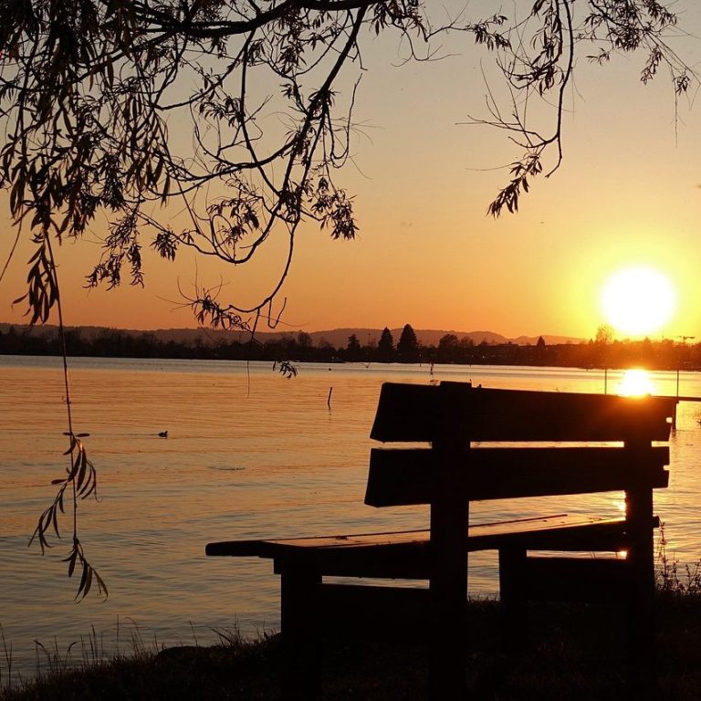 A wooden bench by a lake at sunset, with warm colours reflecting on the water.